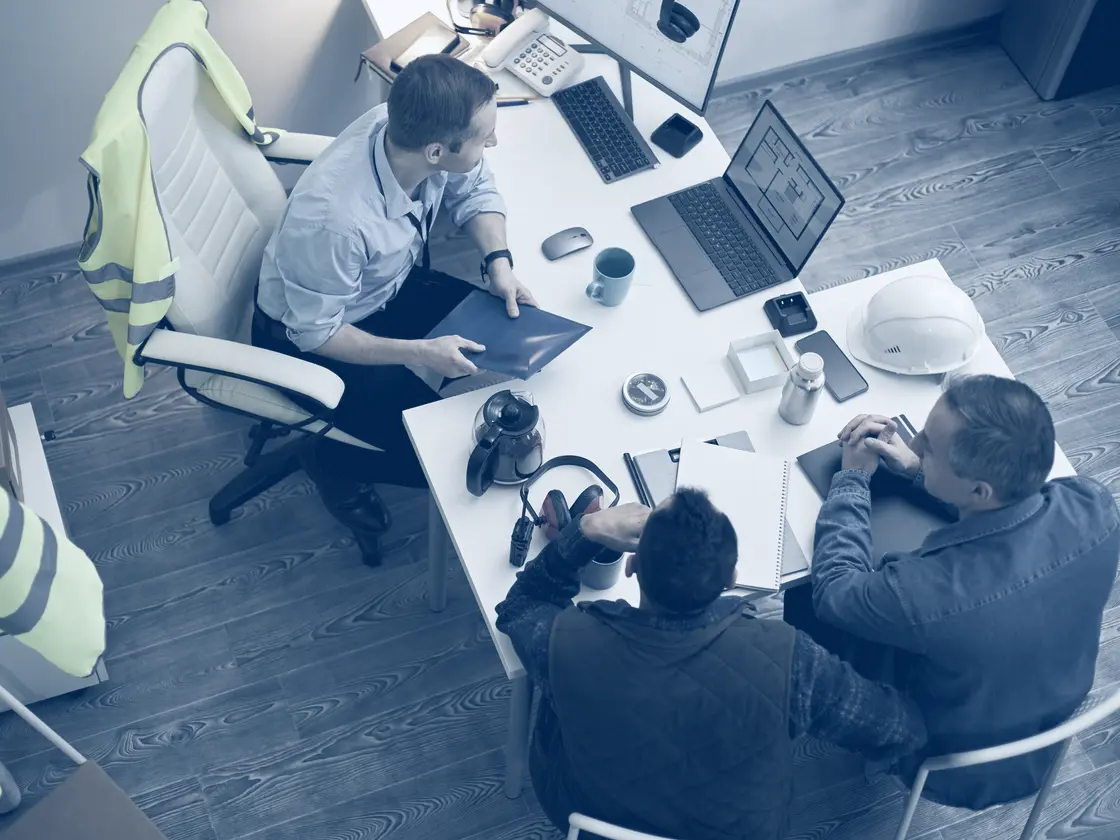 A small team talking around one desk, hi-vis over the chair